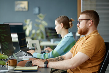 Obraz premium Caucasian young adult man with beard and tattooed arm working on computer next to woman typing at desk in modern office setting, both focused on screens