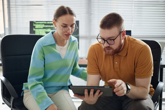 Caucasian young adult man showing something on digital tablet to woman sitting beside him in modern office setting, both focusing on screen together - Powered by Adobe