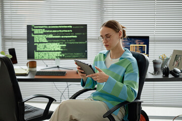 Caucasian young adult woman sitting at desk using digital tablet in modern office, computer...