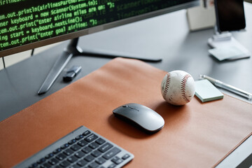 Closeup showing computer keyboard, wireless mouse, baseball, and monitor displaying programming code on desk, illustrating workplace of software developer with sports interest