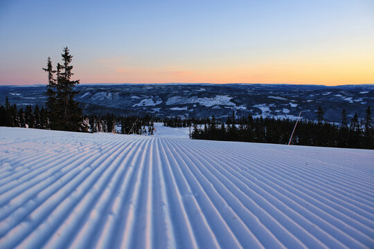 Freshly groomed ski slope at sunrise in Trysil, Norway, with panoramic view of snowy mountains and pine forests. Winter morning alpine landscape.
