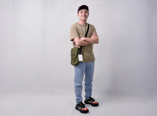 A young man stands with arms crossed, wearing a beige t-shirt, blue jeans, cap, and an olive crossbody bag in studio background © Freddy Chandra