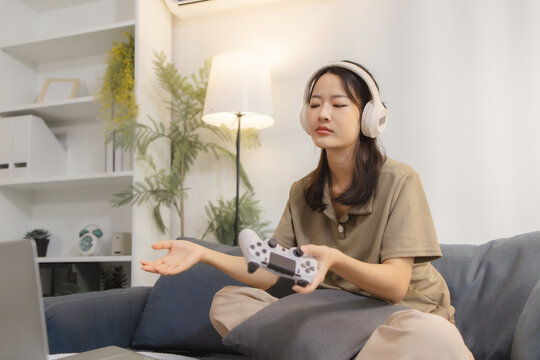 A young woman wearing headphones looks frustrated while playing video games at home. She holds a game controller and reacts with disappointment, showing emotions of stress and failure.