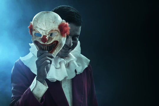 Man hiding behind scary clown mask on dark background, with smoke mist fog, Halloween theme