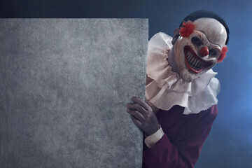Man wearing scary clown mask holding empty blank sign board on dark background with smoke mist fog, Halloween theme