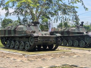 Two camouflaged military tracked vehicles, possibly tanks or armored personnel carriers, displayed outdoors under trees.