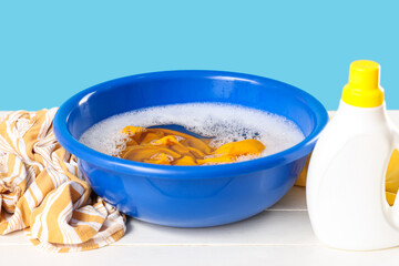 Basin with foam, water, laundry and bottle of detergent on white table against blue background
