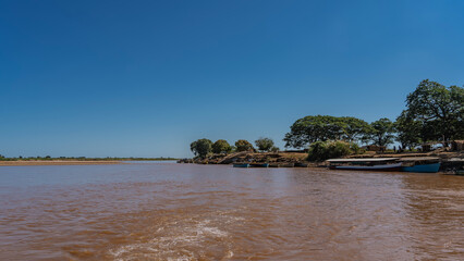 An exotic African river. A foam trail on brown water. The boats are moored at the shore. Green trees on the coast. Clear blue sky. Copy space. Madagascar. Tsiribihina river.