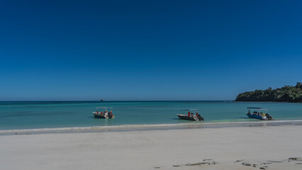 Three motorboats are anchored in the turquoise ocean. Waves are foaming on a white sandy beach without people. A green hill in the distance. Clear blue sky. Copy space. Madagascar. Nosy Iranja 