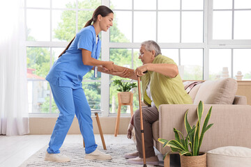 Obraz premium Female caregiver helping happy senior woman to stand up from sofa at home