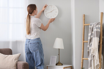 Young woman hanging clock on light wall in living room, back view