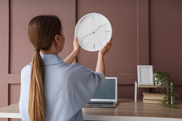 Young woman with wall clock at desk in office, back view