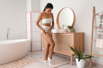 Young African-American woman applying cream to her leg with stretch marks in bathroom