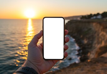 A hand holds a modern smartphone with a blank white screen against a vibrant sunset over the ocean and distant cliffs.