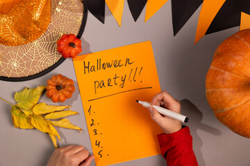 A man writes a list on orange paper with the inscription - Halloween Party next to a pumpkin, miniature pumpkins, autumn leaves and a witch's hat.