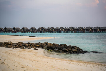 luxury over water villas on stilts stretch across turquoise waters in a tropical resort, with a pristine sandy beach and rocky shoreline in the foreground, under a pastel sky at sunset