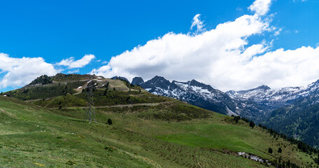 the verdant foothills ascend, they give way to rugged peaks veiled partially by billowing clouds under the vast expanse of a blue sky.