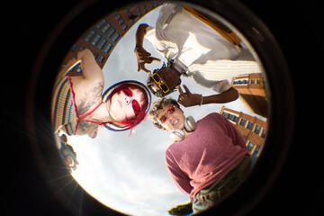 Group of three teenagers, including Caucasian girl with tattoos, Black boy making hand gesture, and hipster boy with headphones, standing outdoors looking down at camera © DragonImages