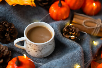 Mug on a blue knit blanket surrounded by tiny pumpkins, pine cones and fairy lights, intimate autumn coffee moment. 