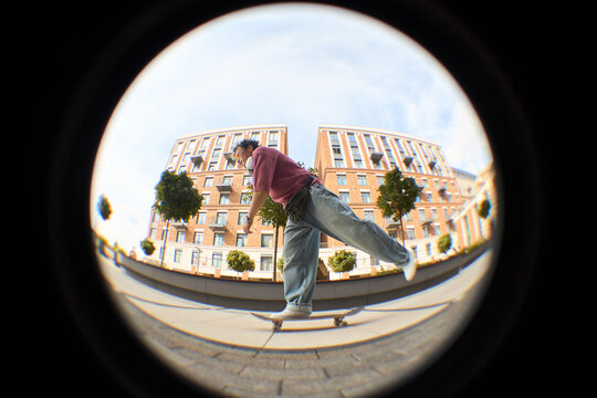 Young adult Caucasian man skateboarding outdoors in urban environment, captured through fisheye lens, wearing casual clothing, balancing on skateboard near modern buildings