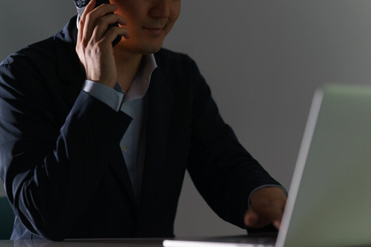 暗い室内でパソコンを操作しながらスマートフォンを使い闇バイトに指示を出す悪徳業者
