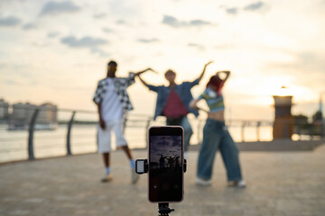 Three multiethnic teenagers dancing outdoors near waterfront, smartphone on tripod recording group, subjects posing with energetic gestures, cityscape and railing in background