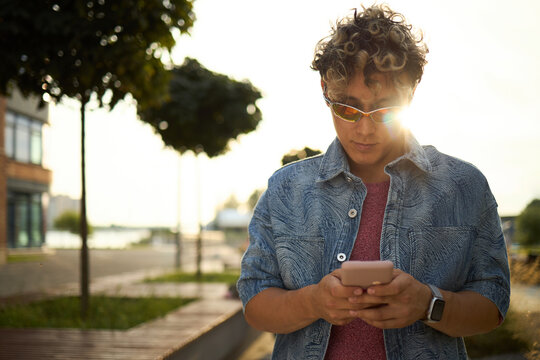 Stylish young Caucasian man standing outdoors using smartphone, wearing sunglasses and smart watch, looking down at device with trees and urban background visible