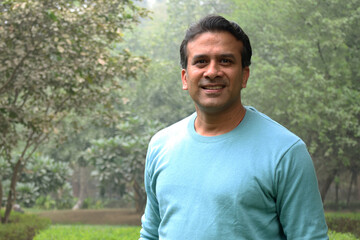 Portrait of a smiling young Indian man standing in casual tshirt in a park, crossing his arms over his chest and looking confidently at the camera.  resting, relaxation feel satisfied glad expressions