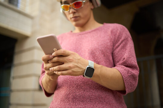 Young Caucasian man wearing headphones and sunglasses using smartphone outdoors, holding device with both hands, smartwatch visible on wrist, standing near building