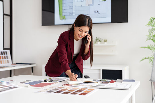 Concentrated young interior designer woman at table with blueprint drawing ,swatches and material , discussing interior design with client  by phone, choosing materials for decorations - Powered by Adobe