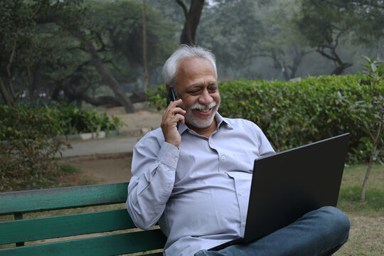 Indian senior man talking on phone while working on laptop at park. Indian Senior man using digital technology at outdoors. Internet banking everywhere
