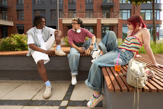 Group of three teenagers, Black, Caucasian male, female, sitting on outdoor bench, talking and relaxing together, headphones and drink cup visible, urban background