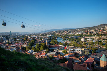 panoramic view of Tbilisi with a cable car over the city, Peace Bridge, Kura River, ancient churches, and modern buildings against mountain backdrop on a clear sunny day © eskstock