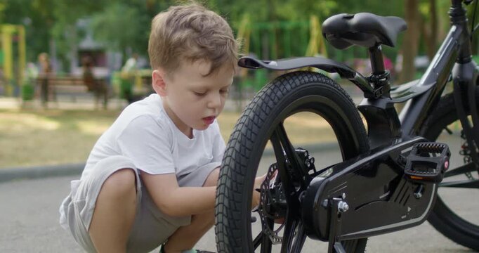 Blond preschooler boy crouches beside bicycle screwing wheel using pliers. Young kid repairs metal part carefully tightening bolt on black metal frame on park path