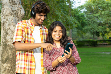 Indian happy couple teenagers friends Student listening to music with wireless earphones, standing in the park. using mobile phone. dressed in casual clothing. Teenagers lifestyle concepts