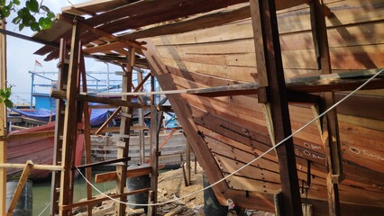Wooden boat being constructed at a shipyard with scaffolding and tools, showcasing traditional craftsmanship and maritime industry in a coastal setting.