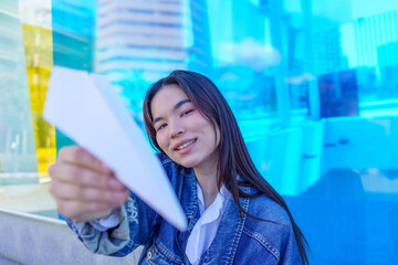 Young adult woman holding a paper airplane in her hand, smiling at the camera outdoors with blue background