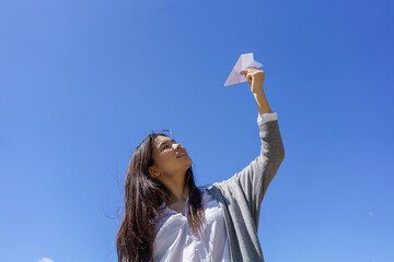 Young woman looking up, smiling, extending arm holding paper airplane against clear blue sky, imagining possibilities and future