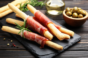 Delicious grissini sticks with prosciutto, peppercorns, rosemary and green olives on wooden table, closeup
