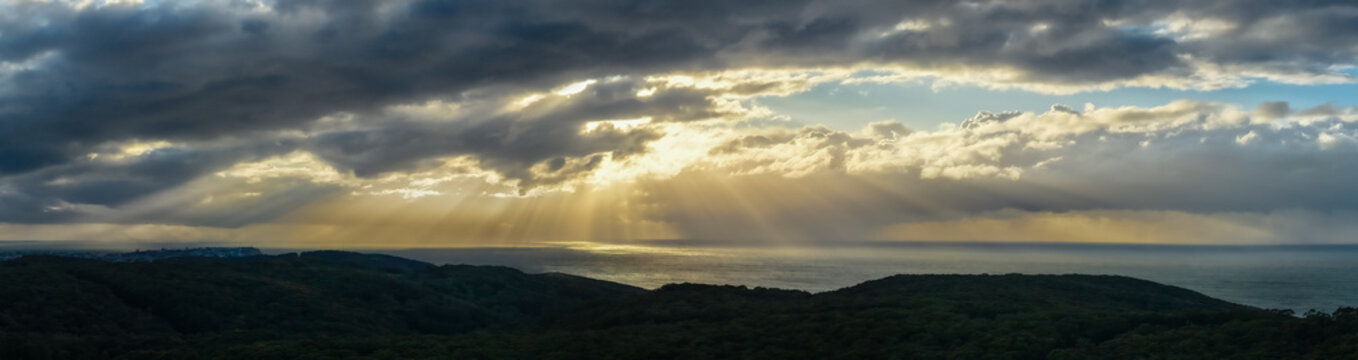 Aerial view of sun rays piercing through dramatic clouds, illuminating the distant ocean beyond the dark, lush landscape, Newcastle, New South Wales, Australia.