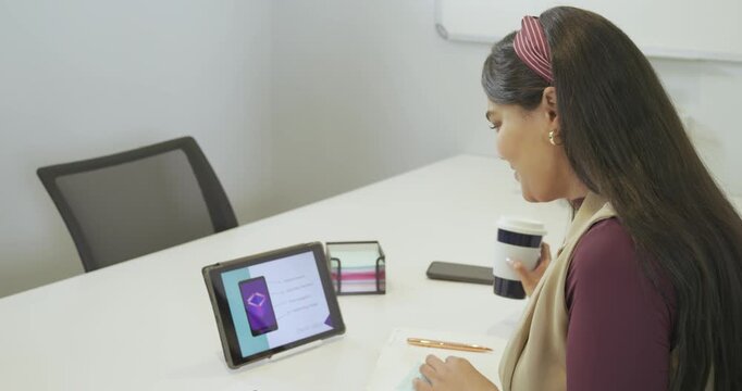 Woman mid-30s reviewing app requirements on tablet at desk, writing design notes and sipping coffee