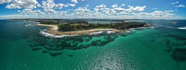 Aerial view of the coastline where turquoise waters meet rugged, rocky shores, and lush greenery stretches towards the horizon, Swansea, New South Wales, Australia.