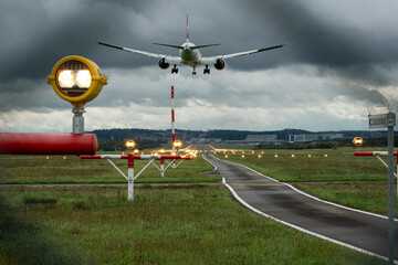 Commercial passenger airplane landing on an illuminated airport runway strip. Ground level, rear of the plane point of view, about to touchdown, telephoto, moody dark clouds, no people