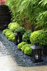 A meticulously landscaped garden bed, featuring dark gray stones, lush green shrubs, and elegant black lanterns, creates a tranquil and serene ambiance.