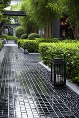 A tranquil, paved walkway winds through a lush, landscaped courtyard, shaded by bamboo and bordered by meticulously trimmed hedges, illuminated by lanterns.