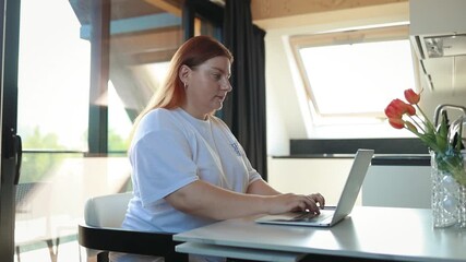 A woman is sitting at a table with a laptop in a bright modern apartment, working remotely from home. On the table, next to her computer, is a glass vase with red tulips, adding a cozy and fresh - Powered by Adobe