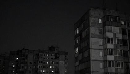 Apartment buildings stand against a dark night sky, with some windows illuminated, conveying a sense of urban living, isolation, and a hint of activity in the darkness.