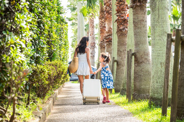 Japanese mother and daughter walking side by side with suitcase through sunny palm tree path during summer family vacation, travel lifestyle with luggage and casual holiday clothing outdoors