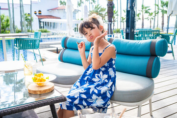 Japanese girl sitting at an outdoor resort café by the pool eating tropical dessert and posing playfully during summer vacation with palm trees bright sunlight and relaxing holiday atmosphere
