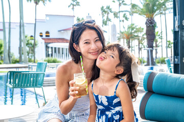 Japanese mother and daughter smiling closely together at a resort poolside café holding a summer drink during tropical vacation with palm trees sunshine and family bonding moment